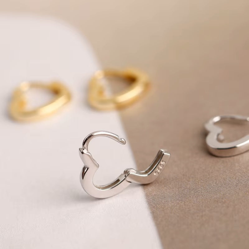 Closed up silver heart hoop earrings in a open position with 925 stamp visible and gold heart hoop earrings blurred on a light background