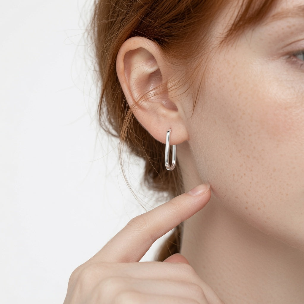 A close-up of a model's ear wearing a small, polished silver hoop earring, with her finger resting near her cheek against a clean white background.