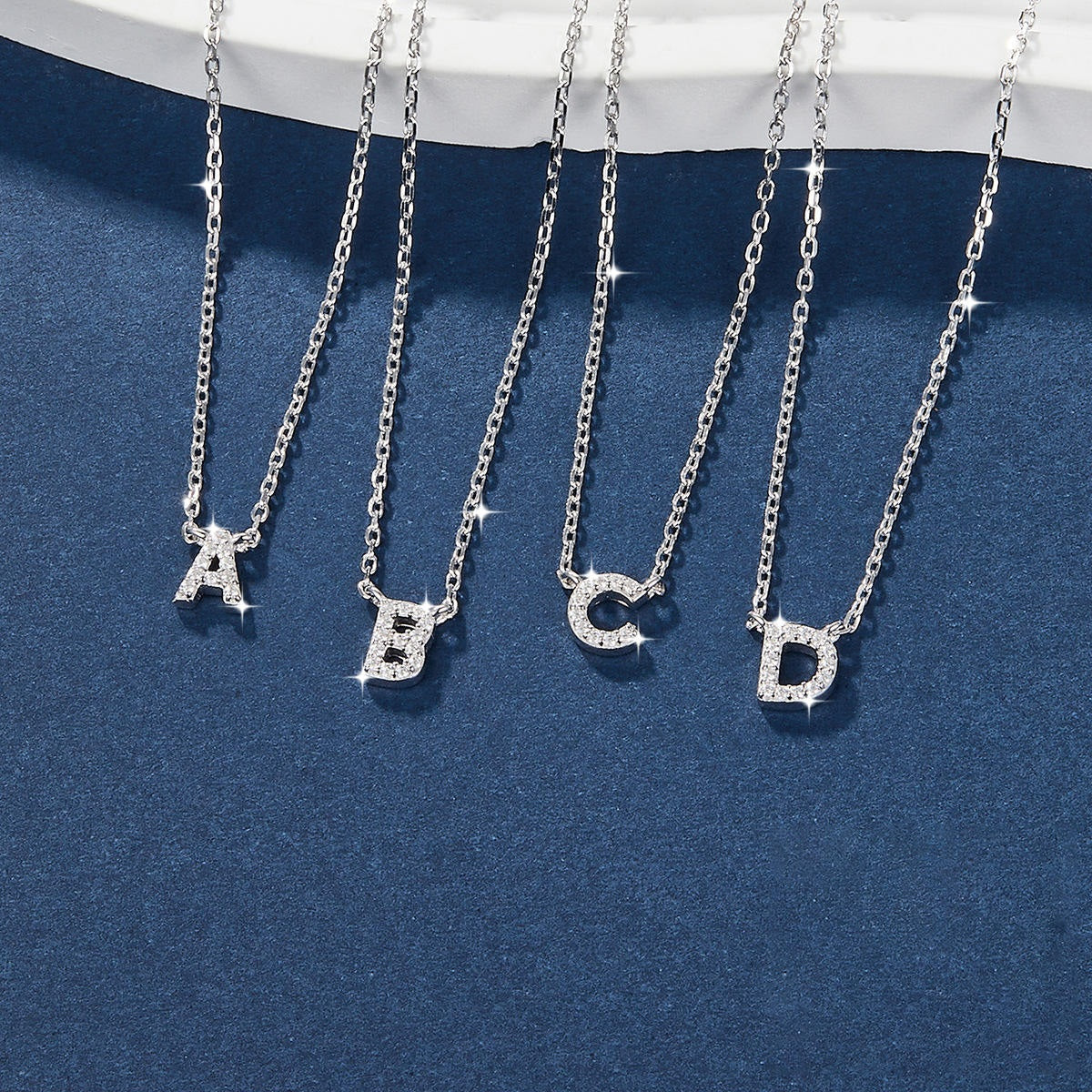 Four silver necklaces with crystal letters A, B, C, D pendants on a blue background